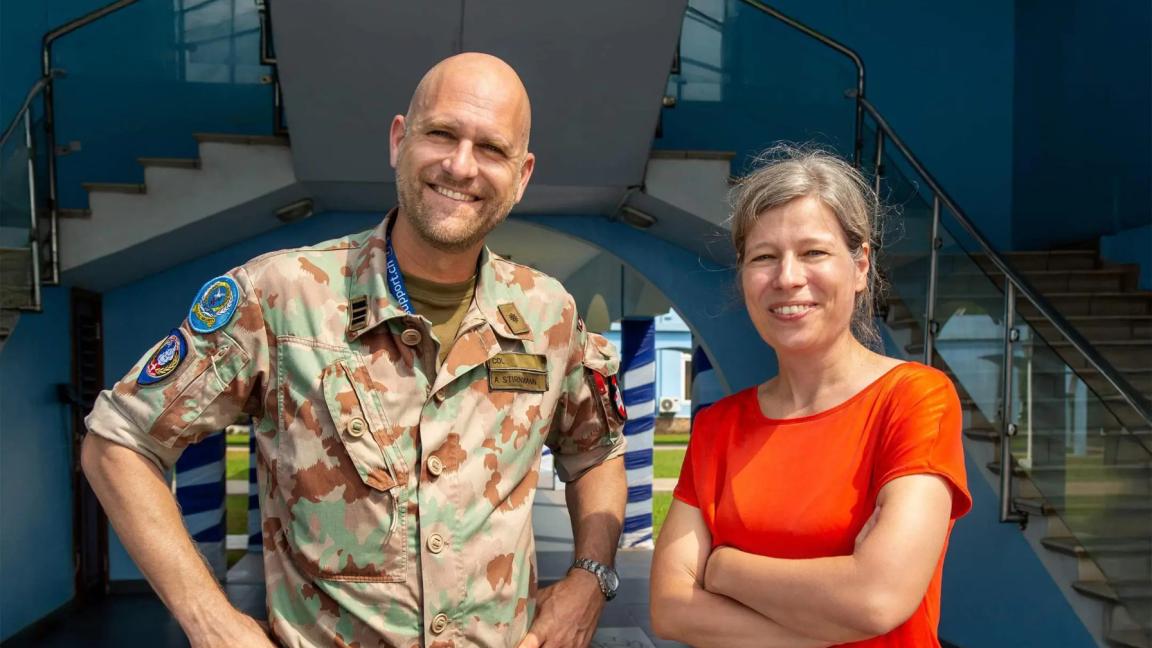 A man in camouflage uniform and a woman in a red shirt smiling in front of a modern staircase.