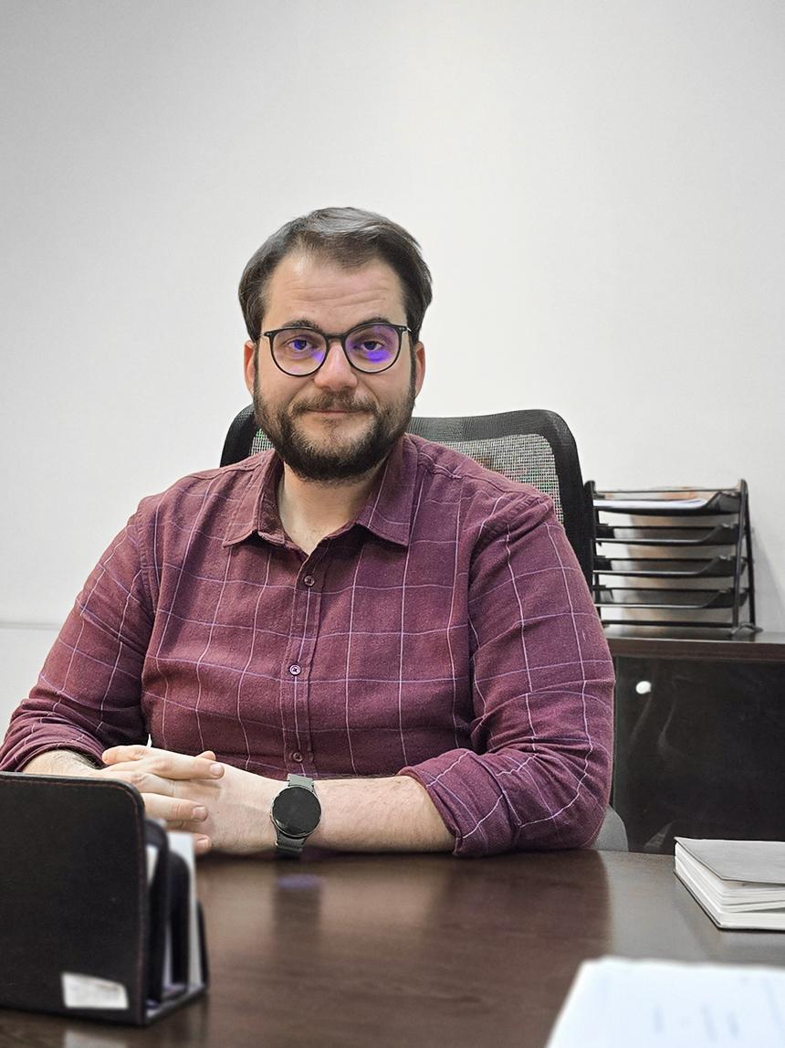 A man wearing glasses and a checkered shirt sits at a desk in an office, smiling at the camera.