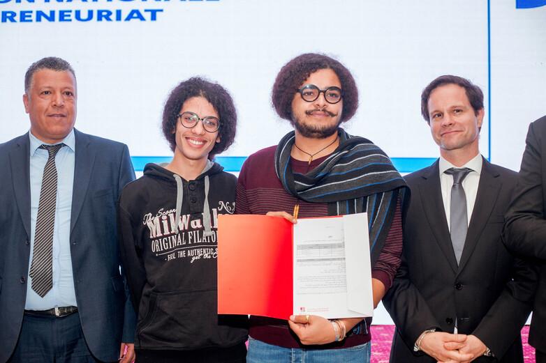 Zwei junge Unternehmer stehen auf einer Bühne und halten eine Auszeichnung des nationalen Unternehmertumwettbewerbs in den Händen.