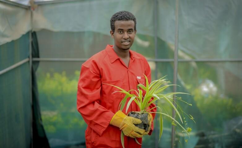 Ein junger Mensch in roter Uniform hält eine Topfpflanze in einem Gewächshaus, als Symbol für umweltfreundliche landwirtschaftliche Praktiken in Somalia.