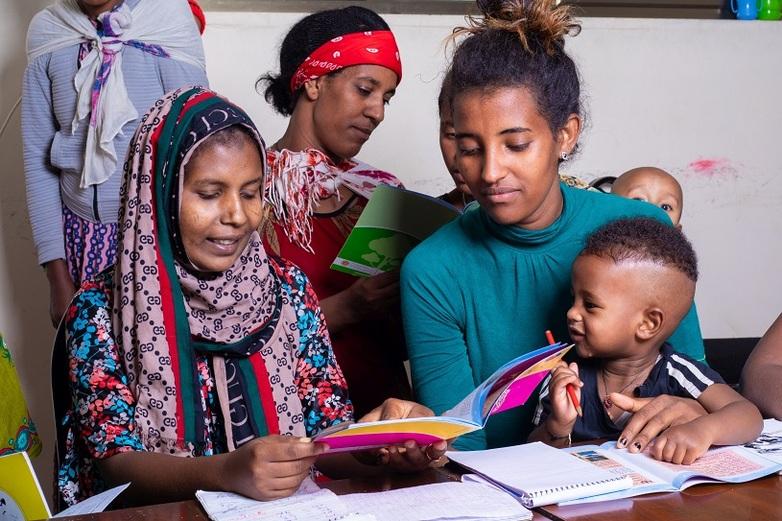 Two women and children fill in workbooks together.