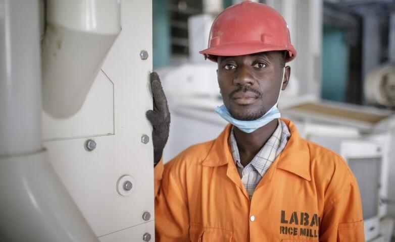 A worker in orange coveralls and hard hat stands in a factory hall.