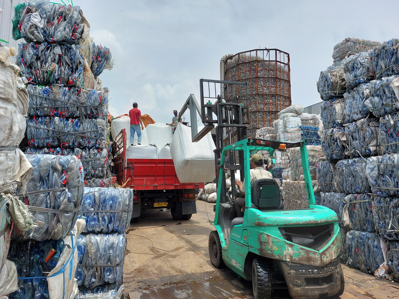 Workers are loading sacks onto a lorry with a forklift truck. Stacks of compressed plastic can be seen on the right and left.