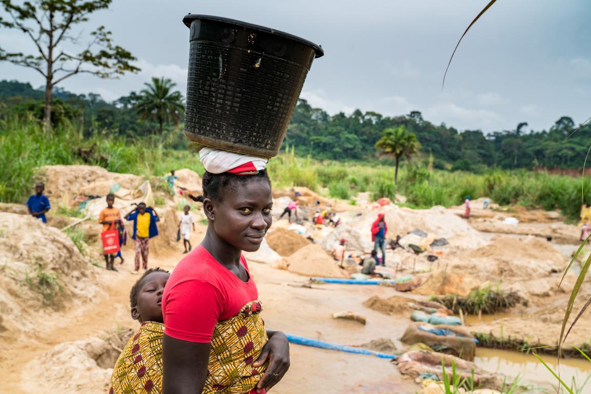 A woman working in the mining sector, who is balancing a bucket on her head and carrying a child on her back.