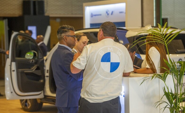 Teilnehmer interagieren an einem BMW-Ausstellungsstand, bei dem ein wasserstoffbetriebenes Fahrzeug präsentiert wird, im Hintergrund eine geöffnete Autotür sowie auf einem Tresen ausgelegtes Werbematerial.