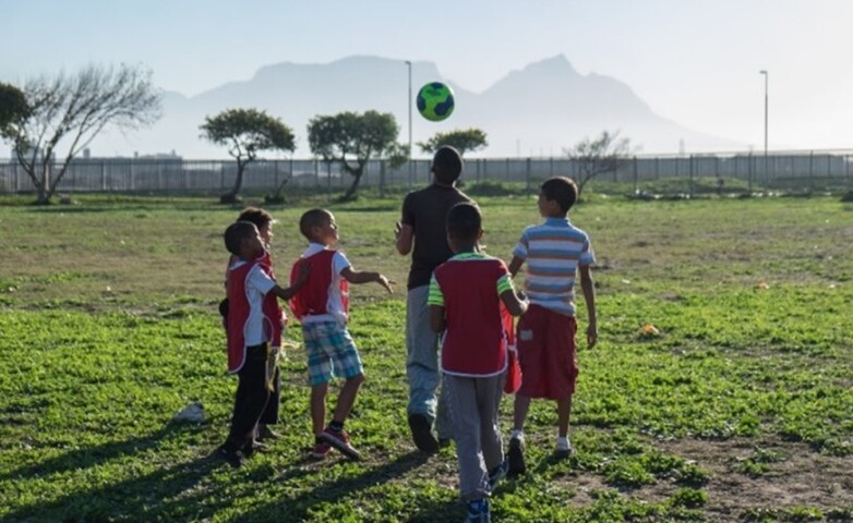 Eine Gruppe von Kindern spielt im Freien vor einem Gebirgshintergrund Fußball.