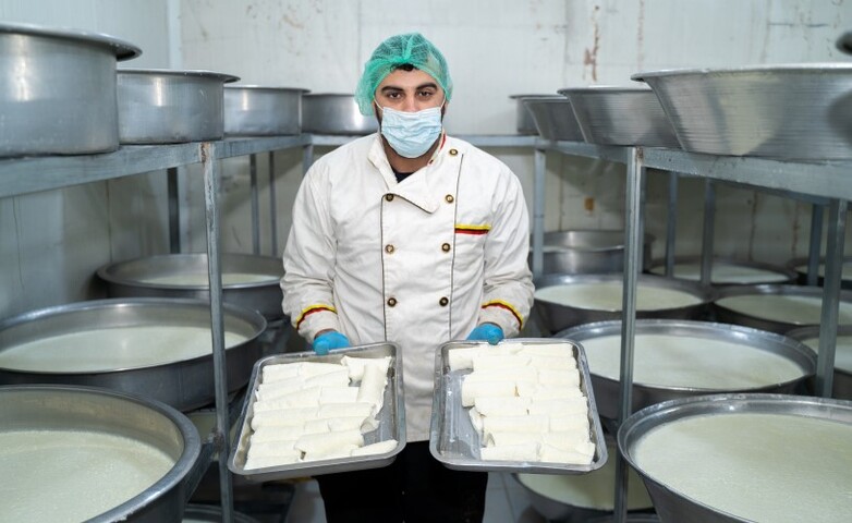 Ein Arbeiter in Hygienekleidung präsentiert frisch zubereiteten Kaymak inmitten mehrerer großer Fässer mit erhitzter Milch in einer Molkerei.