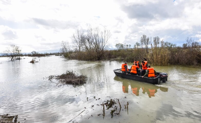 Kleines Boot mit zivilen Rettungskräften in orangefarbenen Schwimmwesten auf dem Weg durch ein Überschwemmungsgebiet in einer ländlichen Region Albaniens.