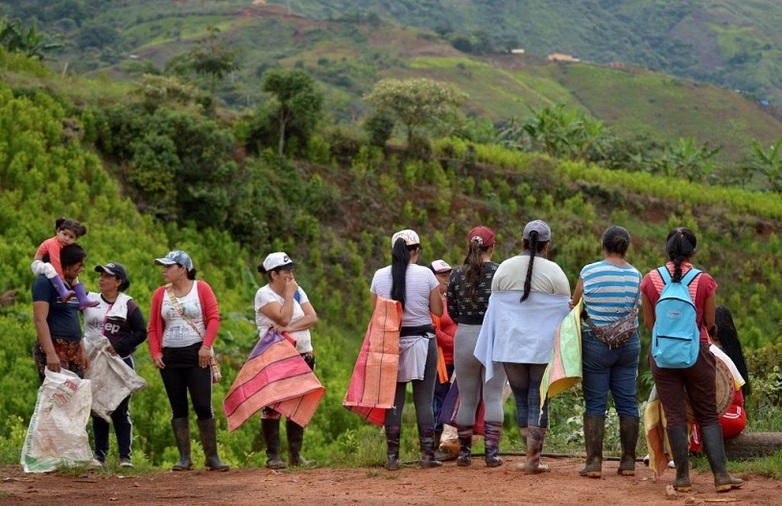 Eine Gruppe von Frauen mit bunten Tüchern und Rucksäcken steht auf einer Lichtung, umgeben von grünen Hügeln und landwirtschaftlicher Vegetation.