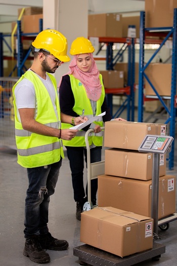 Zwei Studierende stehen in einem Logistiklager mit vielen Paketen und wiegen diese ab.