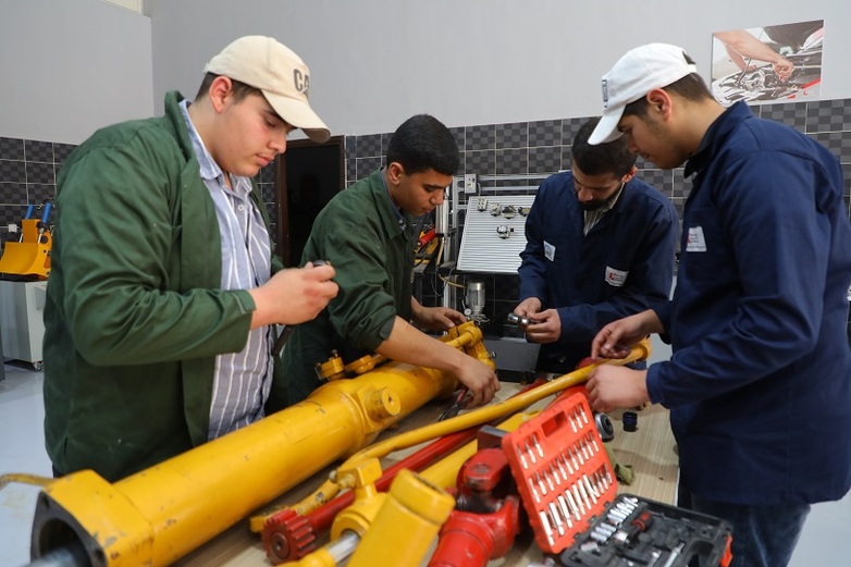 Drei Studenten und ein Ausbilder reparieren ein schweres Maschinenteil in einer praktischen Schulung für Automobiltechnik.