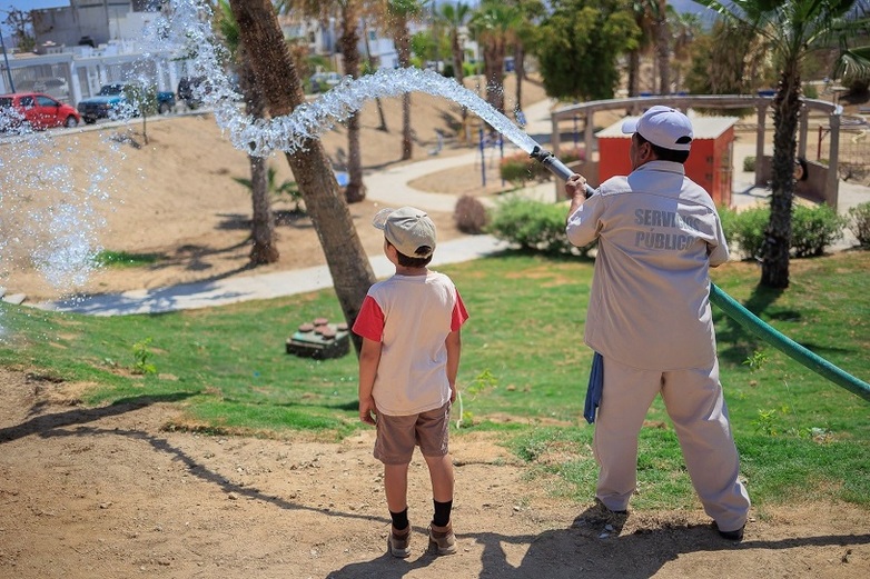 Eine Person bewässert mit einem großen Schlauch einen Stadtpark zur „Cities Challenge Mexico“, während ein Kind zusieht.