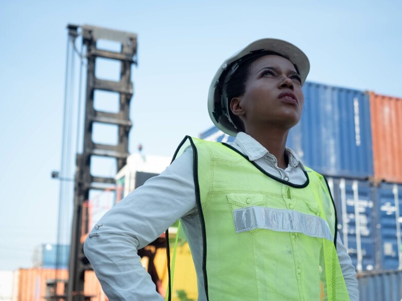 Eine Person in Schutzkleidung schaut in einem Containerhafen nach oben.