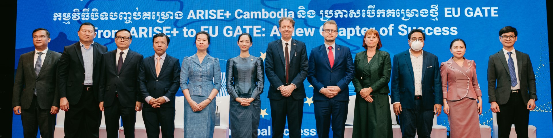 Eine Gruppe von zwölf Personen in formeller Kleidung steht auf der Bühne der EU-GATE-Projektveranstaltung. Im Hintergrund sind Informationen zum Projekt auf Khmer und Englisch zu sehen.