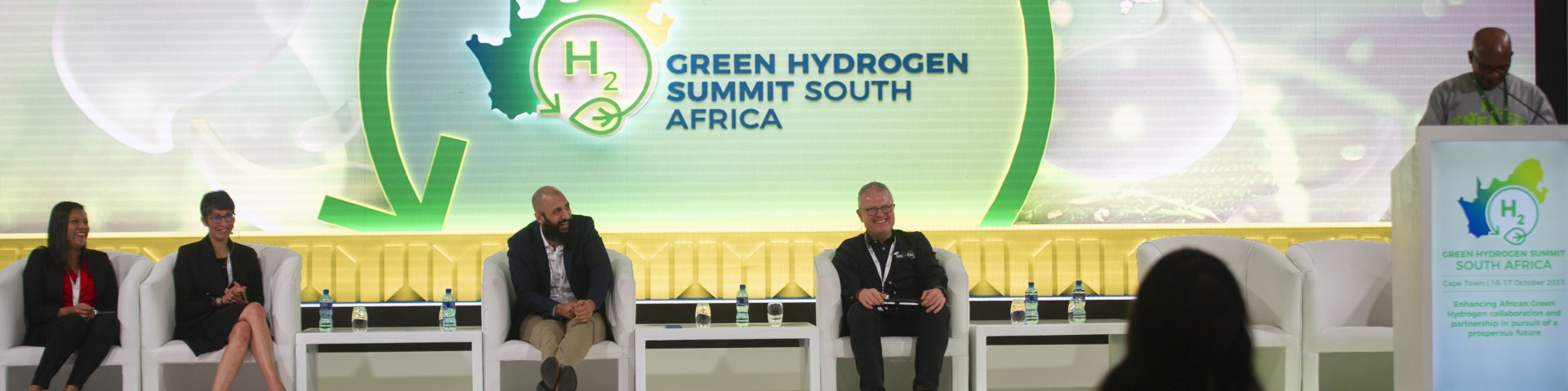 Podiumsdiskussion beim Green Hydrogen Summit in Südafrika mit fünf Teilnehmern auf einer Bühne, das Logo des Gipfels groß im Hintergrund.