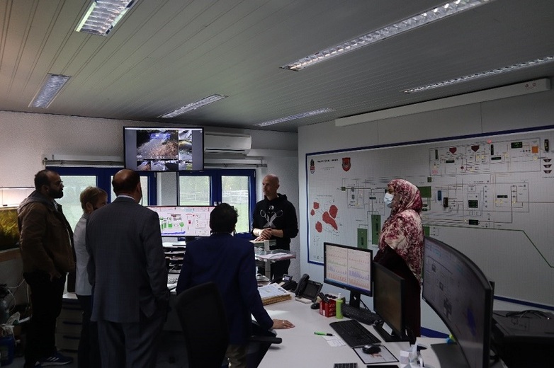 Mehrere Personen stehen in einem Büro mit einem Lageplan an der Wand und schauen auf einen Monitor. 