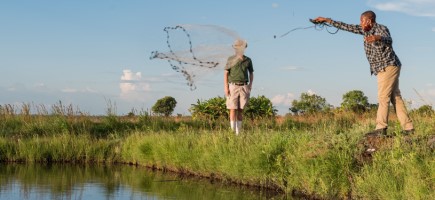 Ein Mann steht an einem Teich und wirft ein Fischernetz aus, im Hintergrund steht ein zweiter Mann.