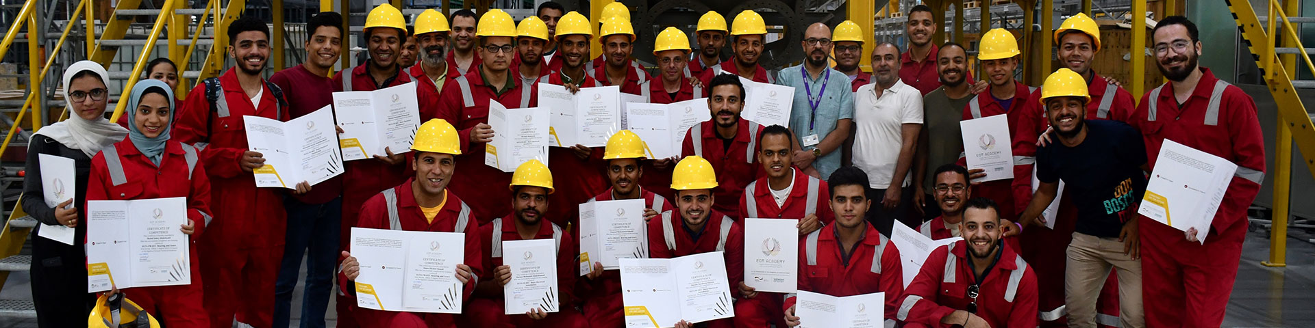 Graduates of a vocational training programme stand in work clothes in a factory. They are holding their certificate in their hand. 