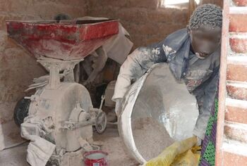 A child covered in white dust uses a large bowl to fill powder into a sack, a grinding machine in the background.