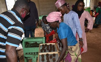 A group of people making briquettes in a workshop with a green machine and wooden trays.