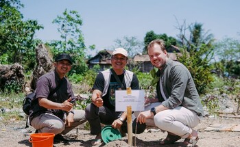 Three men crouching outdoors near a wooden post with a sign, participating in a tree-planting activity in a rural area.