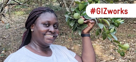 A woman in a white T-shirt stands in front of a tree and smiles. In her left hand she holds a branch of limes. Next to it is the lettering “GIZ works”.