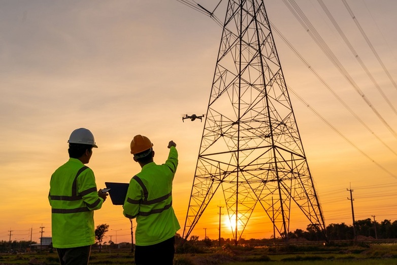 Two engineers in high-visibility jackets and hard hats use a drone to inspect a high-voltage transmission tower at sunset. 