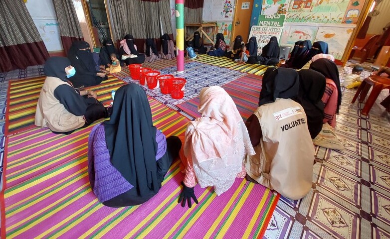 Women in a Rohingya camp participate in an awareness event on gender-based violence, sitting in a circle on colourful mats inside a room decorated with posters.