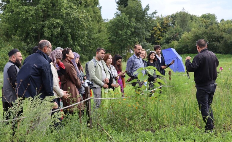 A study group stands in a field, attentively listening to a person speaking, with trees and greenery in the background.