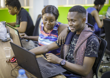 A man and a woman are sitting happily in front of a laptop, in the background a woman is also working on a laptop.