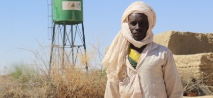 A man – Ibrahim Yatara – standing in front of a green water tank in an arid landscape. 