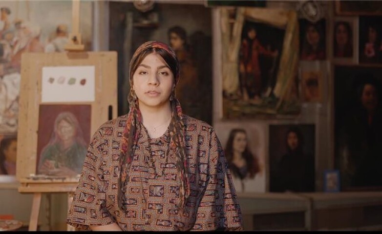 A person sitting in an art studio surrounded by paintings, with an easel displaying a portrait in the foreground.