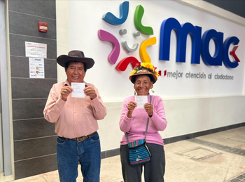 Two people stand in front of the ‘MAC’ Citizen Service Centre and hold their ID cards up to the camera.