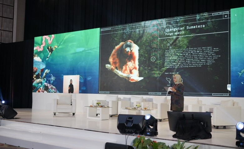 A presenter on stage speaking in front of a large screen displaying an image of a Sumatran orangutan.