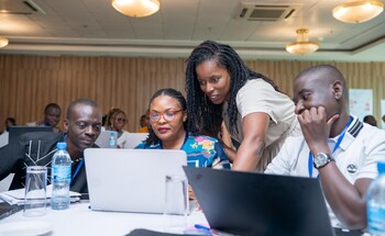 Participants working collaboratively on laptops during a session at the NELGA Research Communication Workshop, with a facilitator providing guidance.