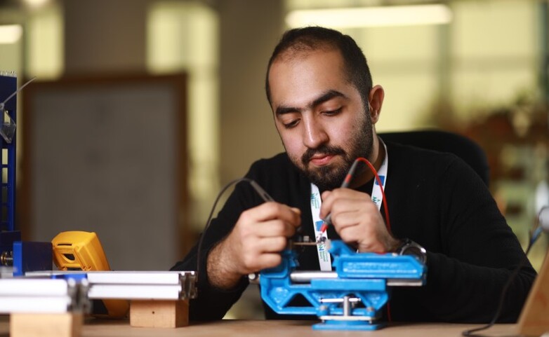 A technician seated at a workstation is using tools to assemble or adjust a piece of blue industrial equipment, with additional tools and machinery visible on the table.