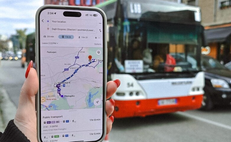 A smartphone displaying a public transport route on Google Maps is held in front of a bus in Tirana, Albania.