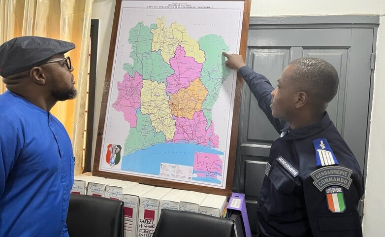 Two individuals discussing a map of Côte d'Ivoire, with one person in a Gendarmerie Commando uniform pointing to specific location bordering Ghana and Burkina Faso.