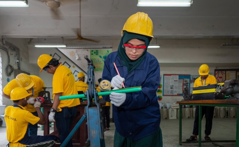 A woman wearing a yellow helmet marks a material for cutting, surrounded by other workers.