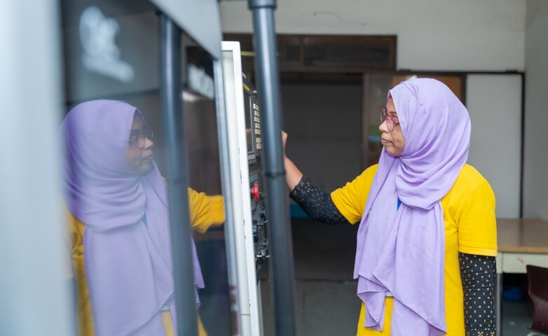 A woman operates a machine, with her reflection visible on the machine’s surface. 