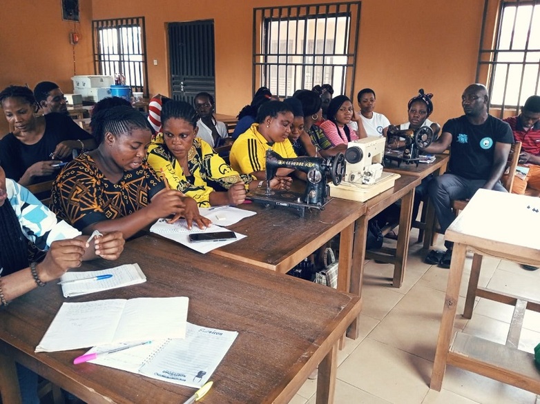 Participants in a tailoring training course sit at tables with sewing machines in front of them.