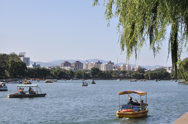 View over a river in Beijing. There are many small boats on the river. Several residential buildings can be seen on the horizon.