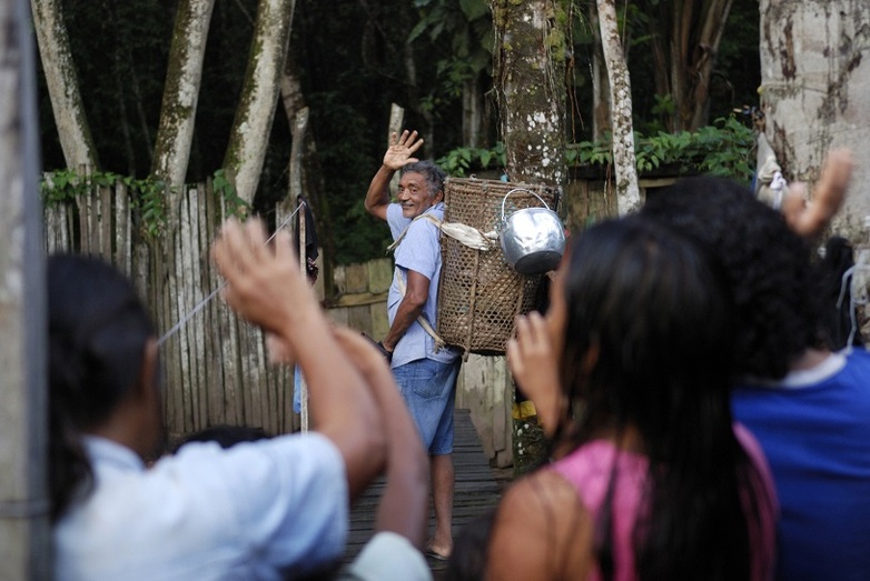 A man carrying a basket on his back stands in front of a forest and waves to a group.