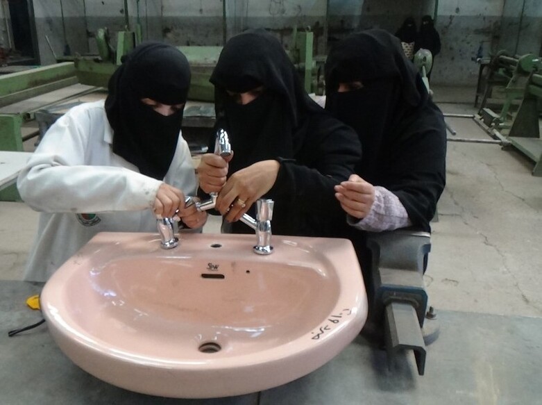 Three women in burqas work at a sink in a factory.