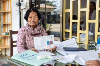 A woman is sitting at a table covered in lists and documents as she registers at a health centre; she is holding up to the camera a laminated equity card featuring her personal details, which entitles her to free medical examinations and treatment.