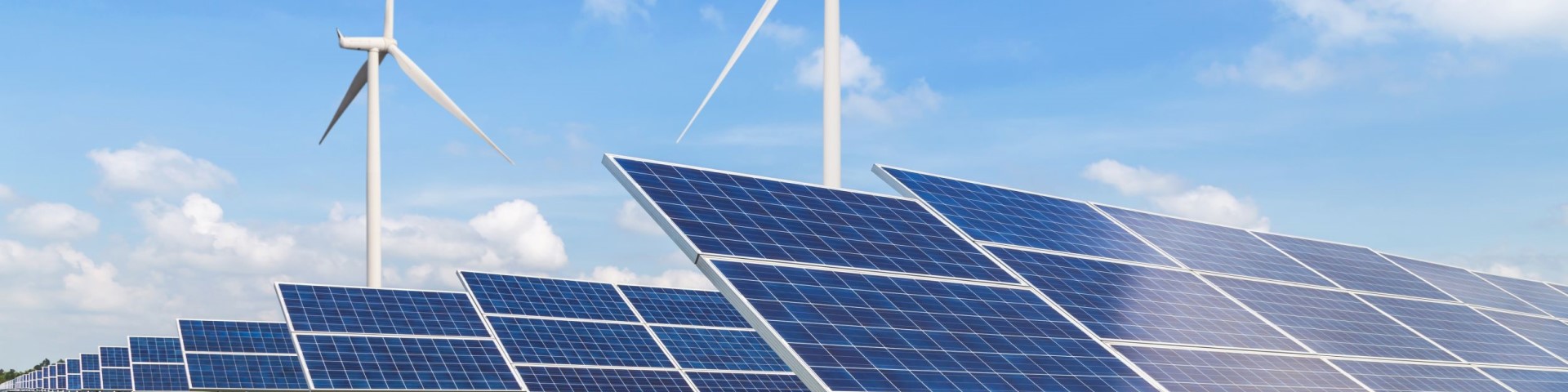 Rows of solar panels in the foreground with wind turbines under a clear blue sky, symbolizing renewable energy integration.