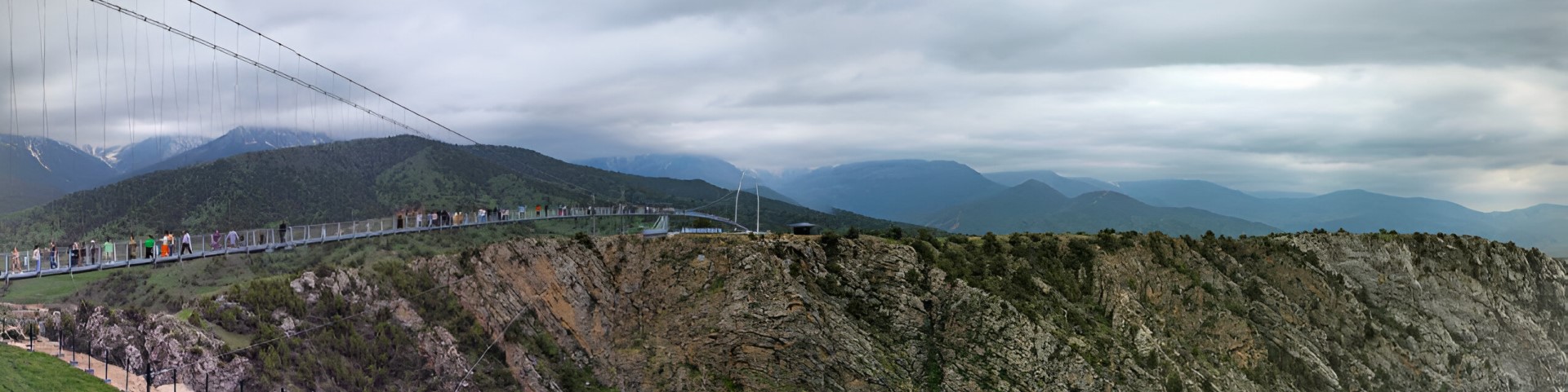 People walk across a modern suspension bridge that crosses a canyon in front of a green landscape.