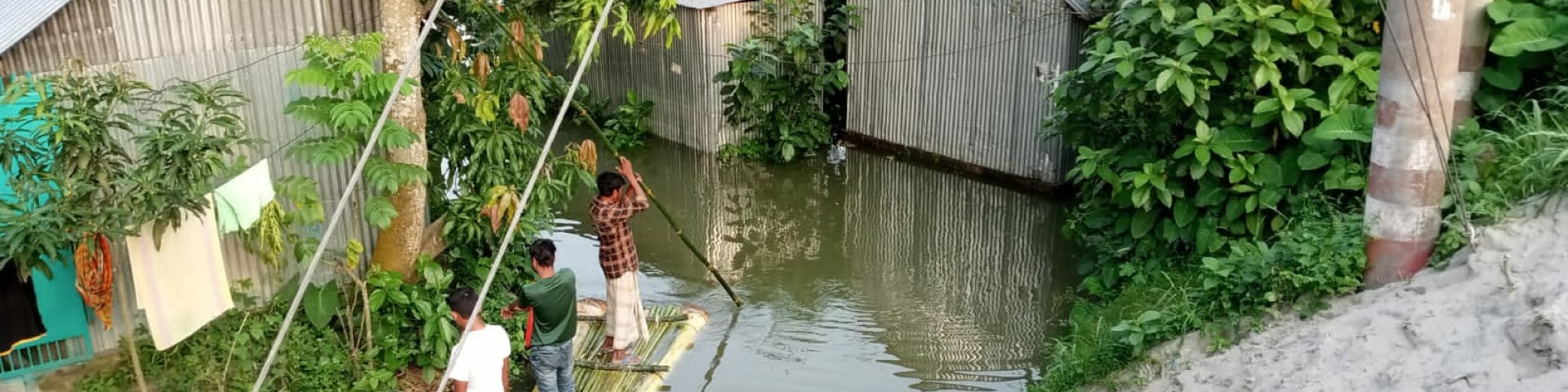 Three people standing on a bamboo raft in a flooded residential area in Bangladesh, surrounded by trees and tin houses.