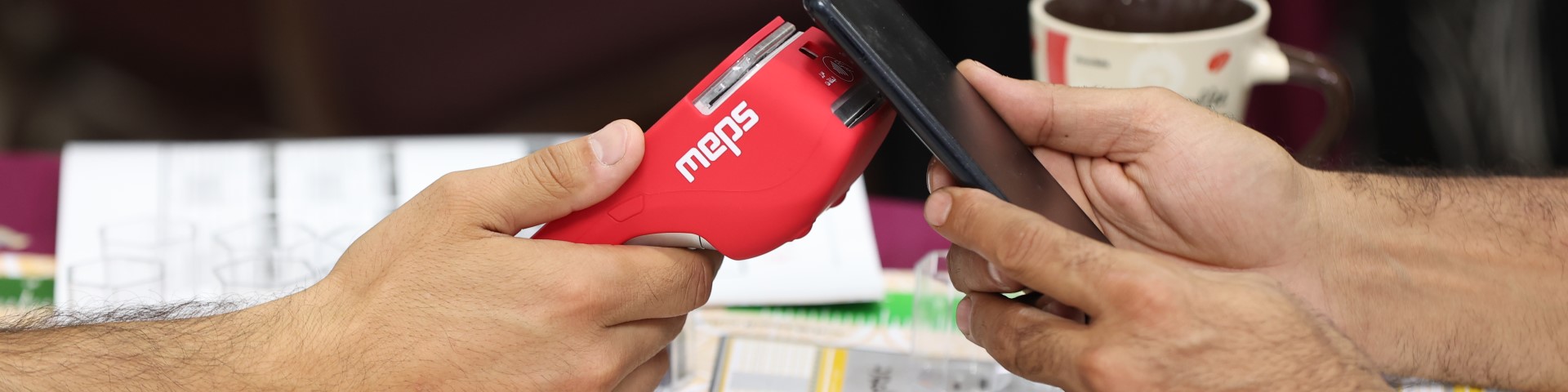 Close-up of two hands holding a mobile phone and a red MEPS payment device, simulating a contactless payment transaction.