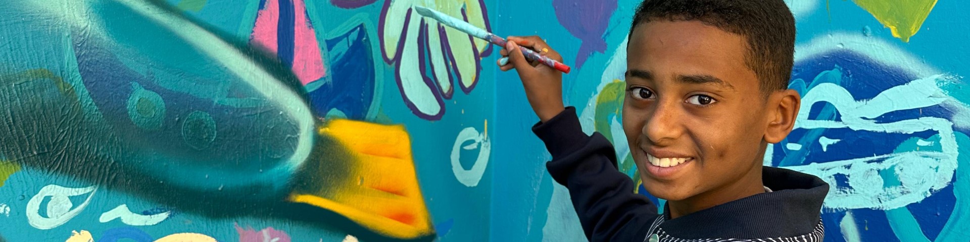 A student paints an underwater scene on a school wall. He smiles and looks into the camera.